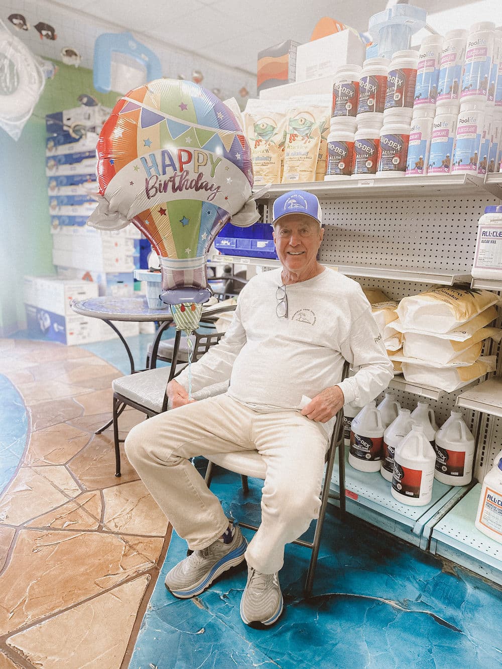 Elderly man sitting in store with "Happy Birthday" balloon and shelves of supplies in background.